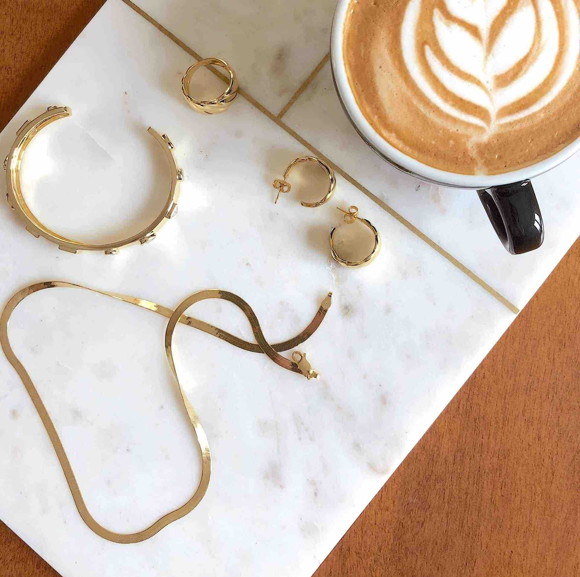 Silber Vergoldete Herringbone Halskette von CANDY mit anderem CANDY Schmuck und einer Tasse Kaffee auf weisser Marmorplatte