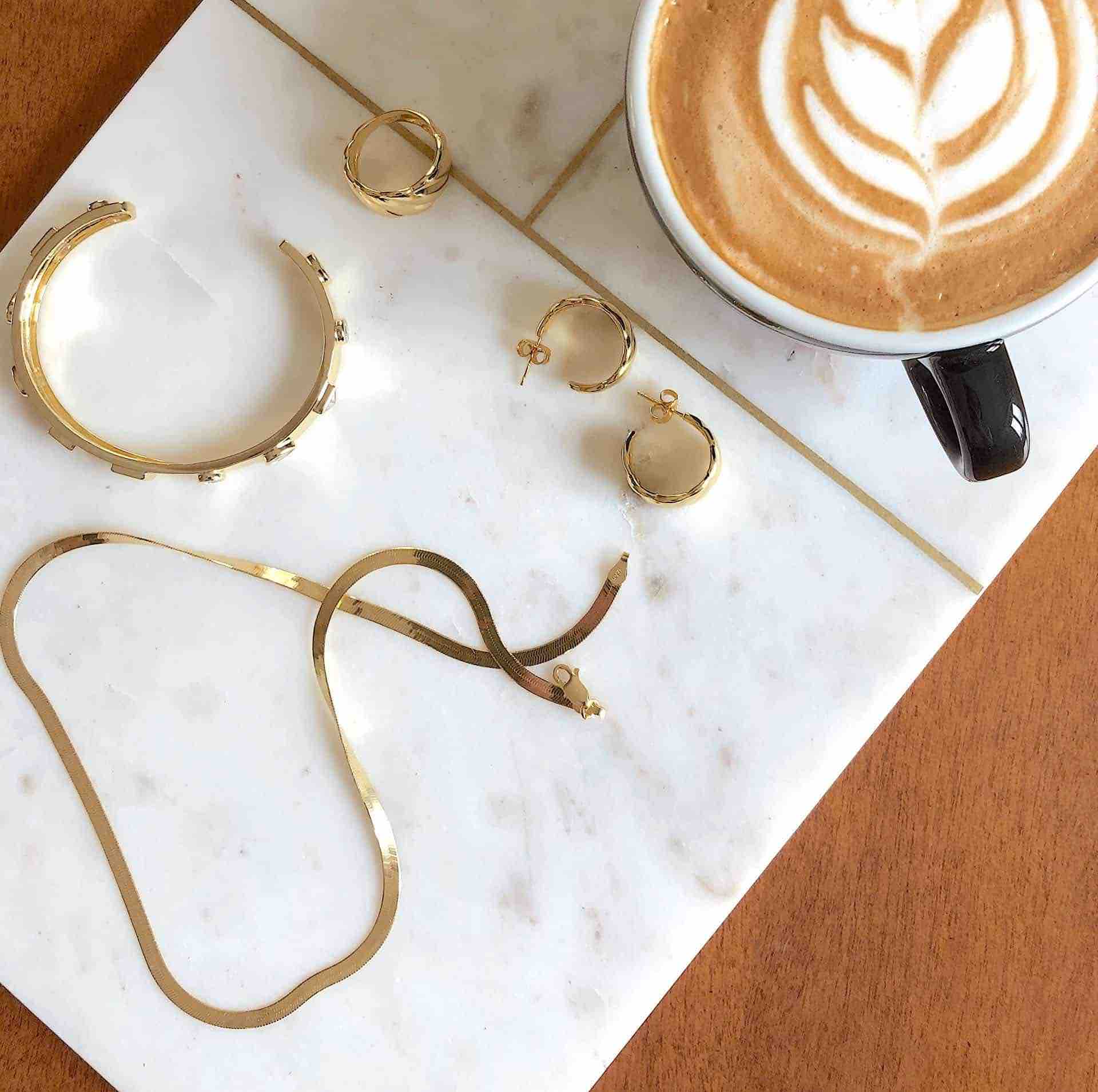 Collar de espiga de plata chapada en oro de CANDY con otras joyas de CANDY y una taza de café sobre una losa de mármol blanco